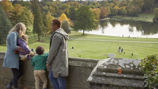 A family of four standing on a stone terrace that overlooks a lake bordered with trees in autumn colours of yellows and browns